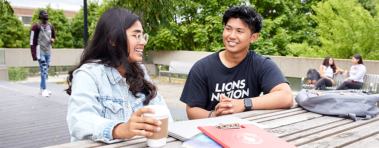 two students sitting on a bench with books in front while they smile and talk