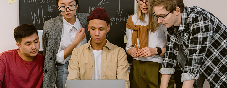 five people in a group look at a laptop screen while one of them types on the keyboard