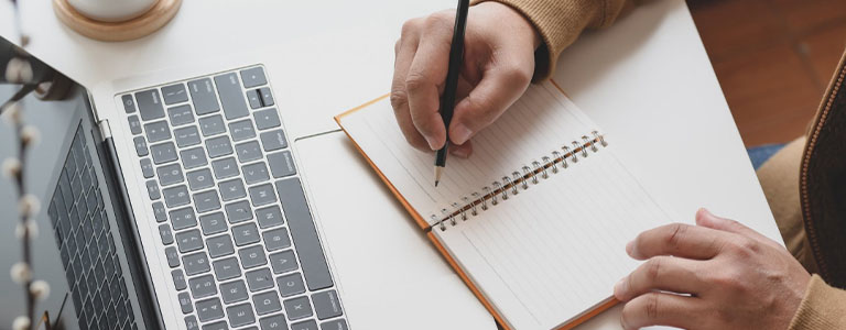 person takes notes while reading a laptop screen.