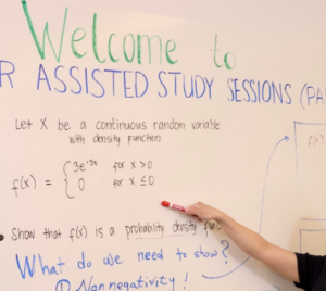 A photo of a peer-assisted study session. A student is at the front of the whiteboard pointing to text written on the board.