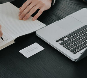 person takes notes with pen and paper while looking at computer screen