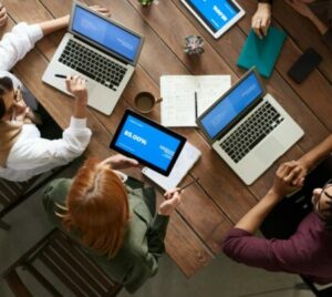 top down view of people talking together and working at a table around laptops, tablets, and notebooks.